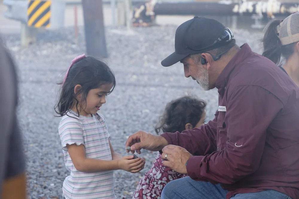 Adult crouching down to show a young girl something small at the waterfront