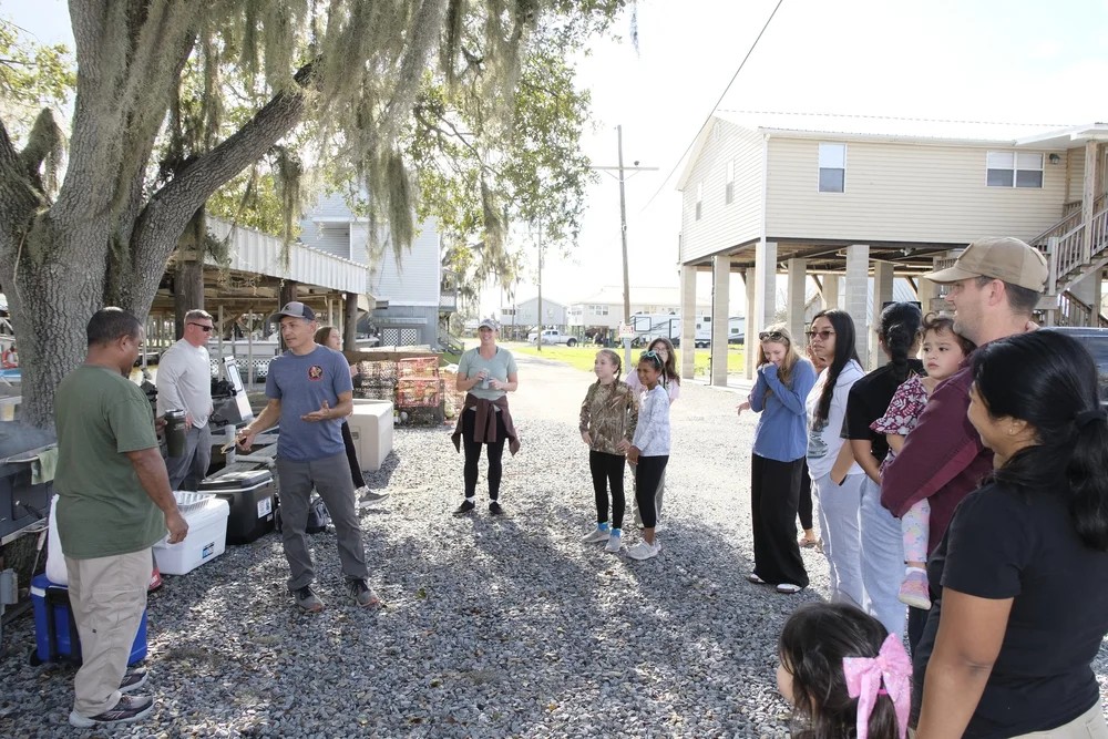 Club leader addressing a large group of families under Spanish moss trees at the bayou